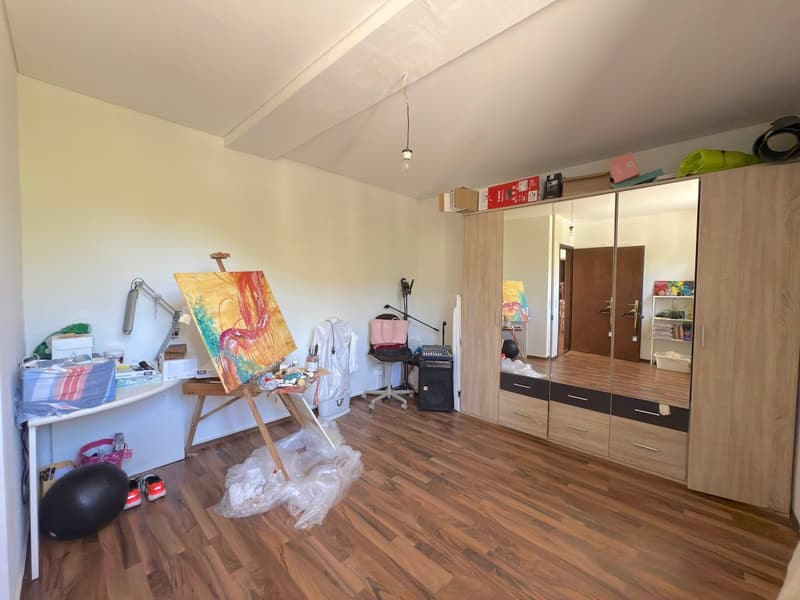 Spacious room with hardwood flooring, white walls, and a large mirrored wardrobe. An easel with a colorful painting and art supplies are set up, indicating this is likely an artist's studio or workspace. The room also has a desk and shelves with various i