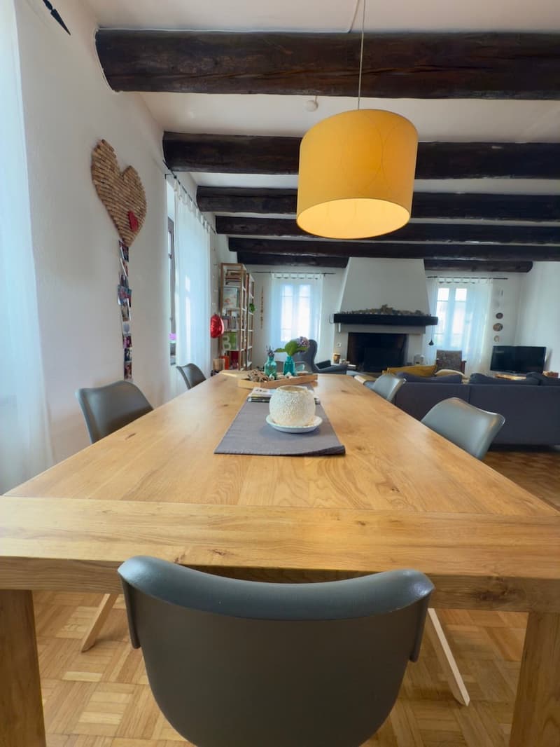 Spacious dining area with a large wooden table, grey chairs, and a yellow pendant light. The room features exposed wooden beams on the ceiling and a fireplace in the background.