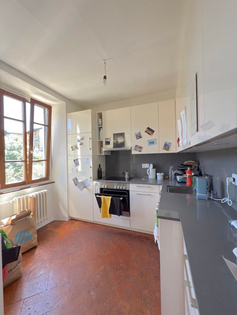 Spacious kitchen with white cabinets, gray countertops, stove, oven, refrigerator, and various kitchen appliances and accessories. The room has large windows providing natural light and a tiled floor.