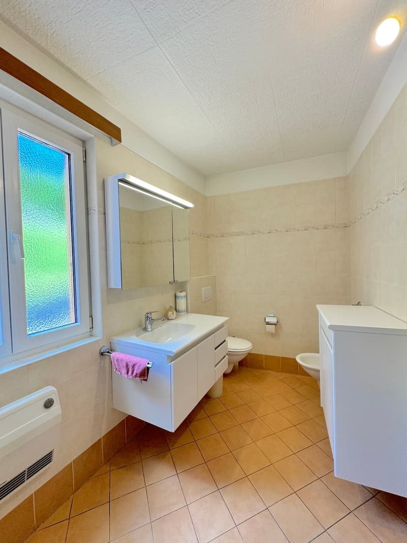 Modern bathroom with tiled floor, white cabinet, mirror with light, sink with faucet, toilet, bidet, and a window with colorful glass.