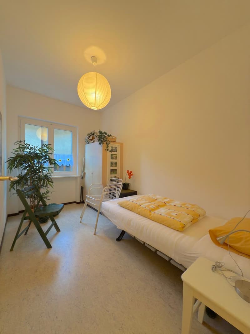 small bedroom, white walls, single bed with yellow pillow, white nightstand, white lamp, small window, wooden chair, wooden folding chair, cabinet, flower vase, potted plant