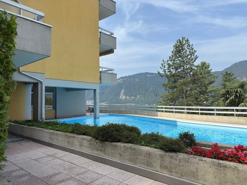 Two-story building with balconies, large swimming pool with landscaped area, mountainous view in the distance