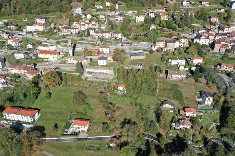 Aerial view of the village with several buildings and houses, green fields, and trees.