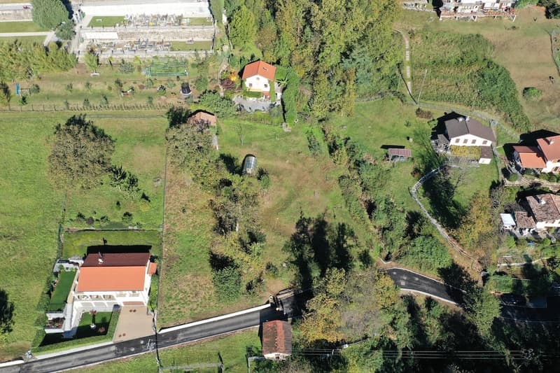 Aerial view of several houses and a plot of land in a countryside area, surrounded by trees and grass, with a road passing by.