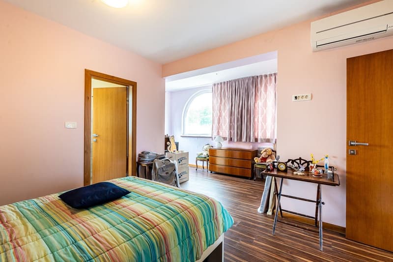 Bedroom with bed, wooden floor, pink walls, wooden door, air conditioner, window with curtains, wooden cabinet, table with items, and storage boxes.