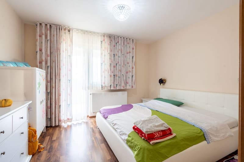 Bedroom with bed, white headboard, white wardrobe, wooden floor, white curtains, white radiator, ceiling lamp, yellow backpack, green pillow
