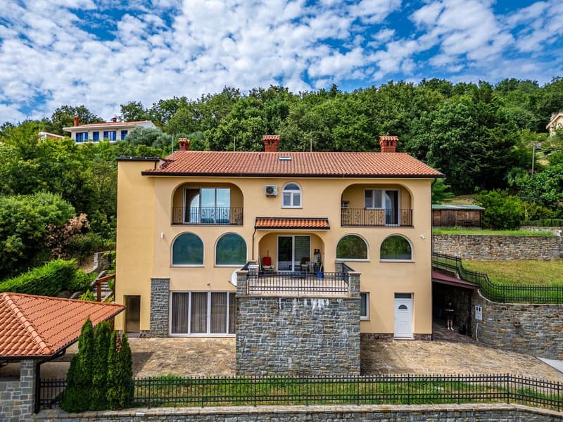 Two-story house, yellow exterior, red roof, multiple windows, balcony, stone wall, fenced yard, greenery