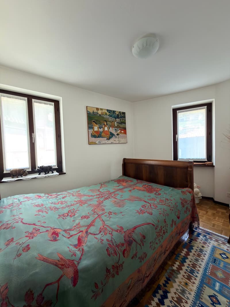 Bedroom with bed, wooden headboard, floral bedspread, two windows, painting on wall, wooden floor, rug, ceiling lamp