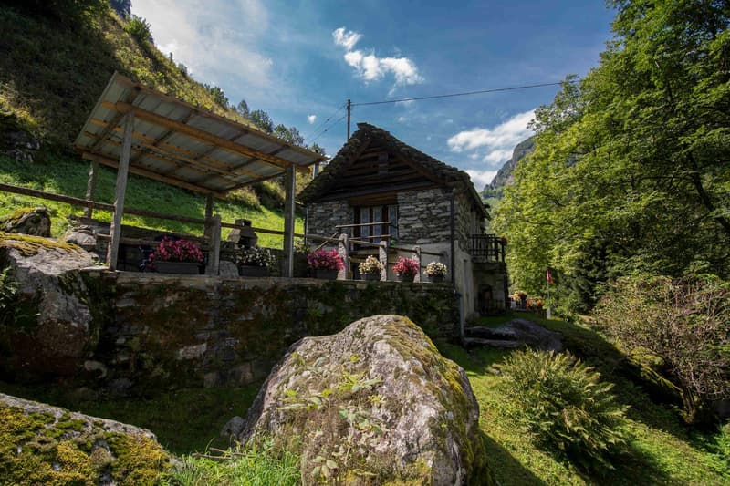 Stone cottage with wooden balcony, surrounded by greenery and mountains