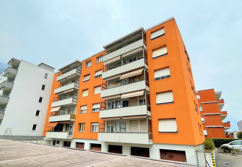 Multi-story apartment building with orange and white exterior, balconies, and parking spaces on the ground level.
