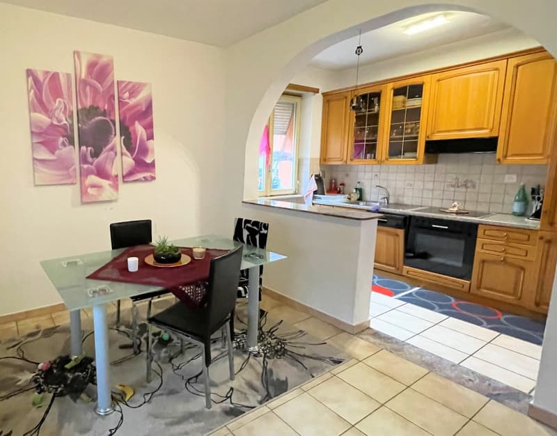 The image shows a combined kitchen and dining room area. The kitchen features wooden cabinets and a tiled backsplash. A dishwasher is visible in the kitchen. The dining area has a glass-topped table with black chairs. The walls are decorated with large fl