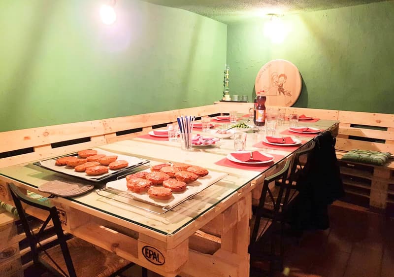 A dining table set up with multiple dishes and drinks for a gathering, wooden benches on the sides, green walls