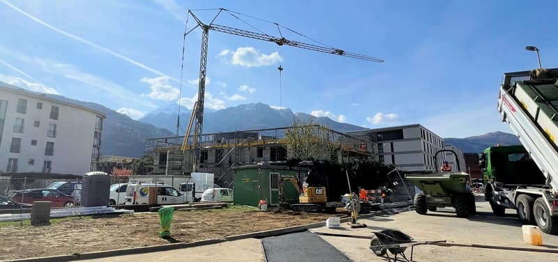 An ongoing construction site with multiple machinery vehicles, workers, a large crane, and nearby buildings, all under a clear blue sky.