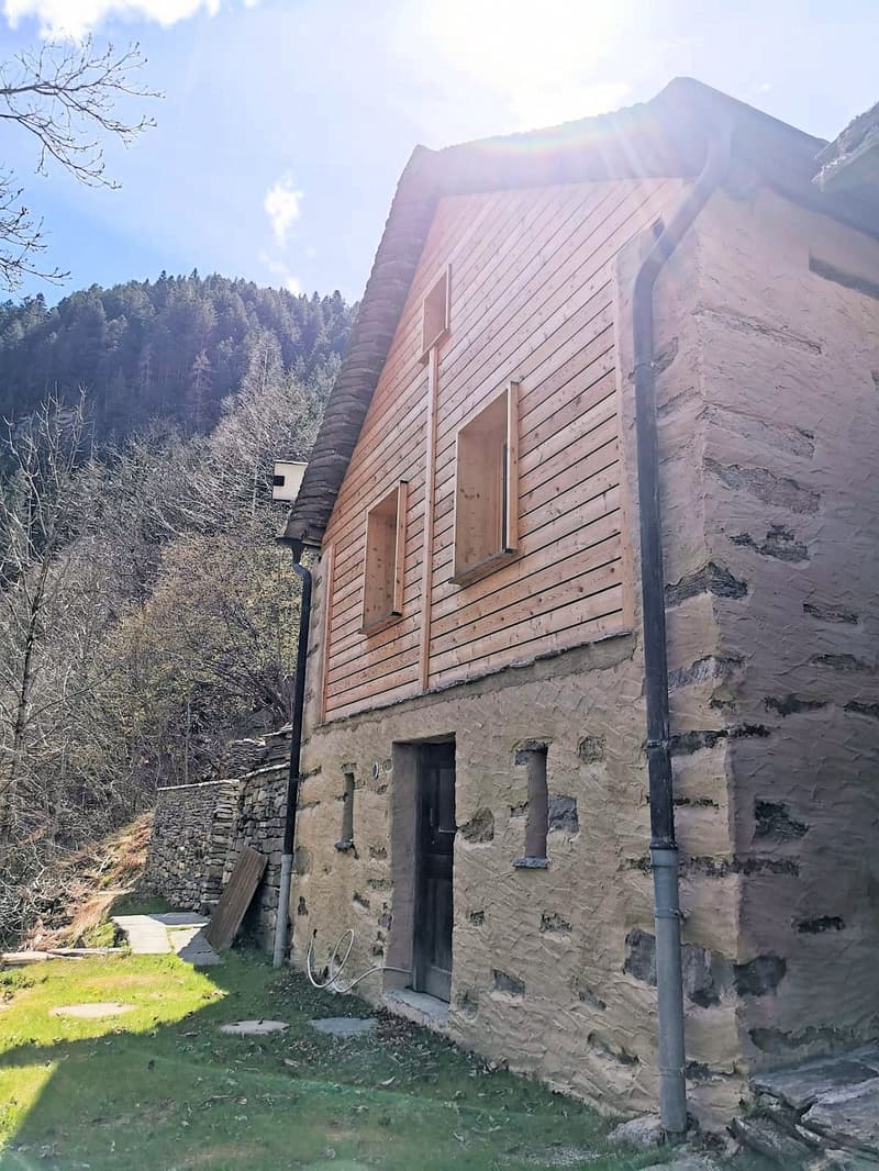 A rustic house with wooden sidings, a stone wall, a roof with a chimney and a wooden entrance.