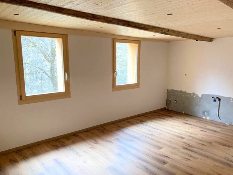 Empty room, wooden floor, wooden ceiling beam, two windows with view of trees, white walls, exposed brick wall