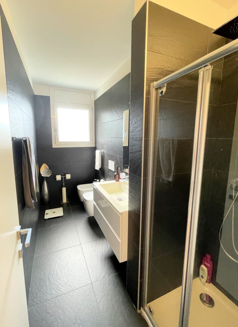 A modern bathroom featuring a white sink with a faucet, a wall-mounted toilet, a glass shower enclosure, and black tiled flooring.