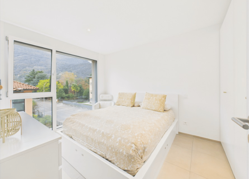 Modern bedroom with a double bed, white walls, and beige bedding. Two large windows allow for a view of the mountains. The room includes a headboard and a nightstand with a decorative object.