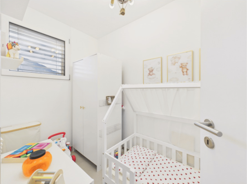 Child's room with white walls, wooden desk, double bunk bed, toys, pictures, white wardrobe, lamp, window with blinds