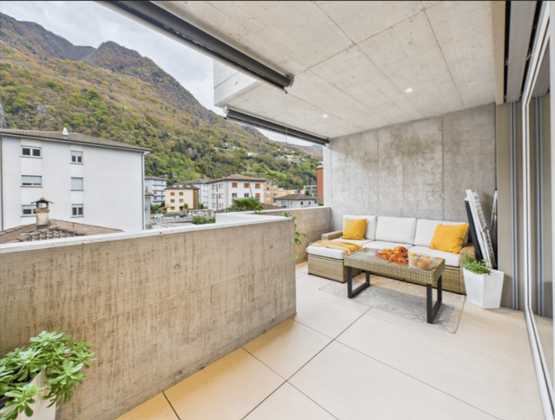 Modern furnished balcony with beige tiles, a white sofa, a coffee table with flowers, white planters, and a view of a mountain