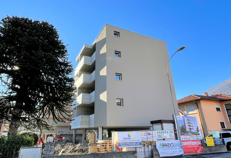 Newly constructed apartment building with multiple floors, concrete walls, and balconies. It has an elevated ground floor and is surrounded by a fence, trees, and construction vehicles.