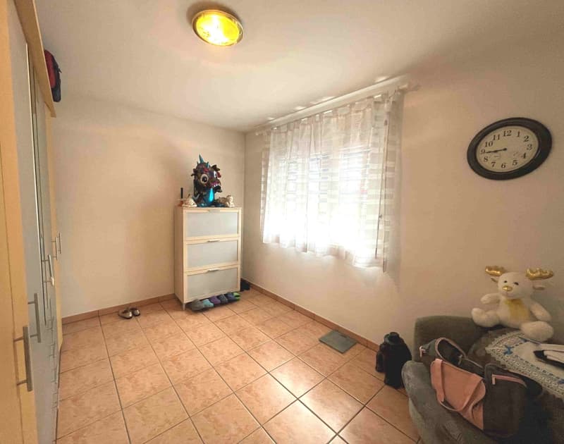 Small room with tiled floor, white walls, ceiling lamp, cabinet, clock, drawers, and a window with curtains. A sofa, a plush toy, bags, and shoes are also present.