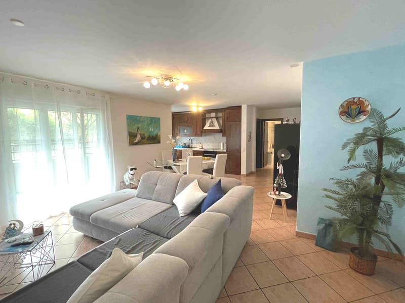 Large L-shaped sofa, white and blue cushions, glass table, round side table, potted plant, open view of kitchen with dining table and chairs