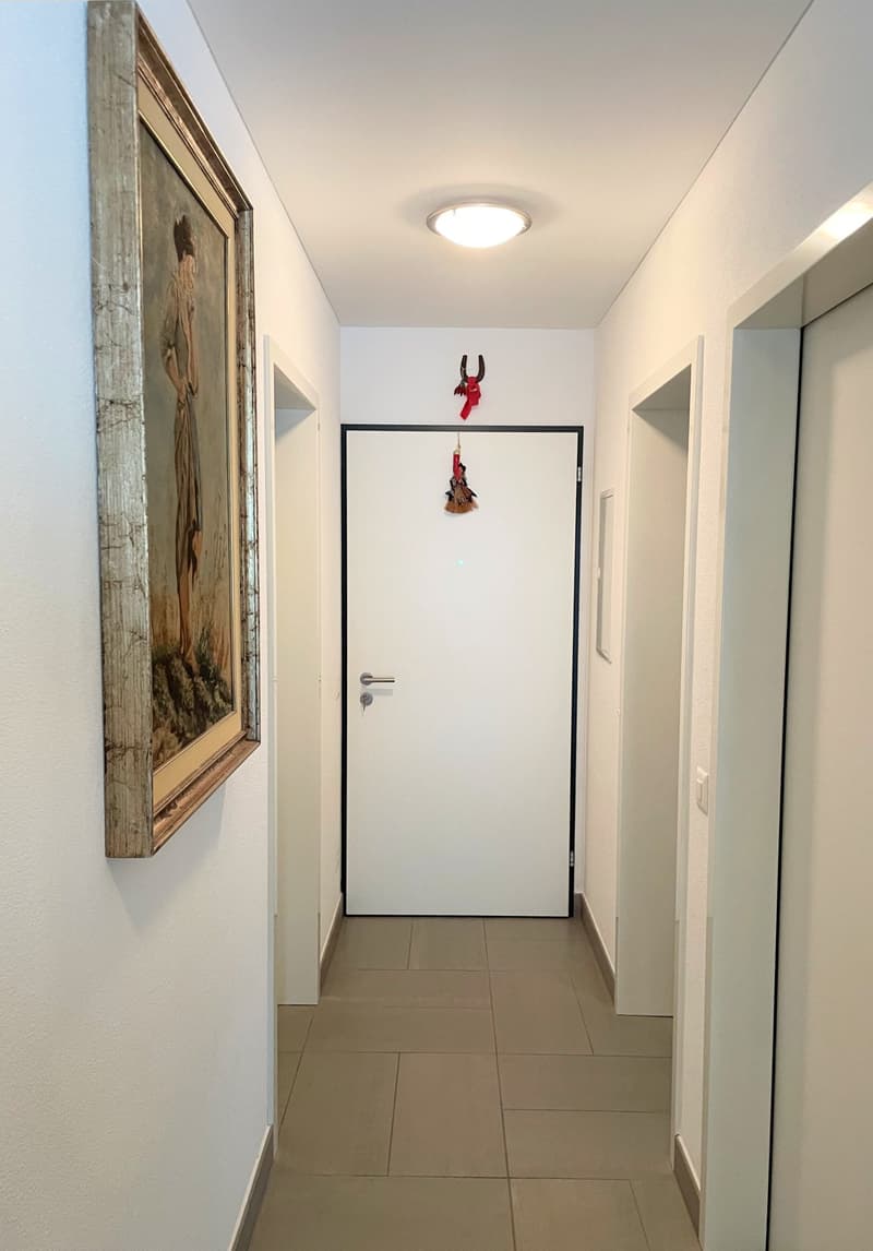White walls, tiled floor, an entrance door, a picture frame on the left wall, and a small ornament hanging above the door.