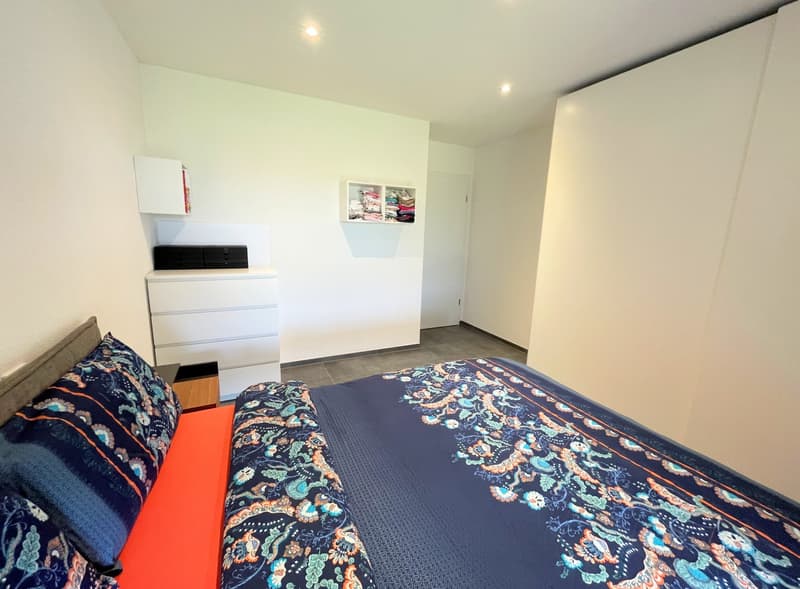 Bedroom with bed, pillows, and a white dresser with drawers. Wall-mounted shelves with folded clothes. White walls and tiled floor.