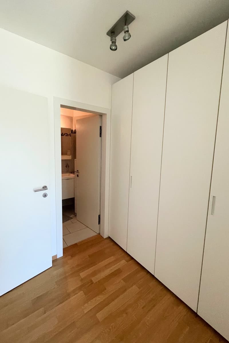 Empty room with wooden floor, white walls, white door, white cabinets, and a bathroom visible through the open door.