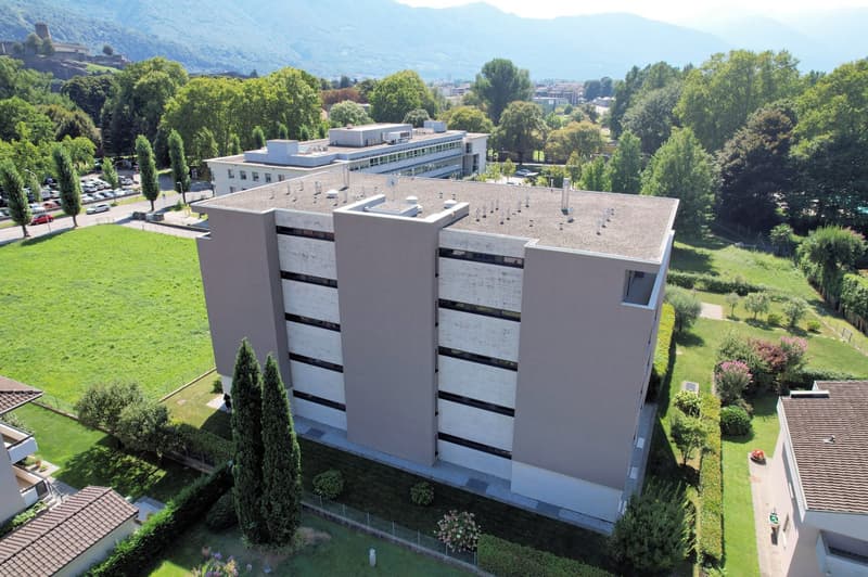 Aerial view of a large building with multiple floors, surrounded by trees and grass, with a parking lot and other buildings in the background.