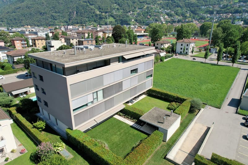 Modern apartment building with multiple floors, surrounded by greenery and a large lawn, with a distant view of mountains and a city skyline.