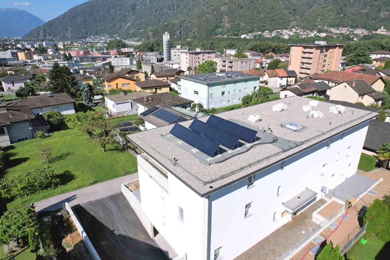 Aerial view of a white building with solar panels on the roof, surrounded by other buildings and trees, with a mountain in the background.