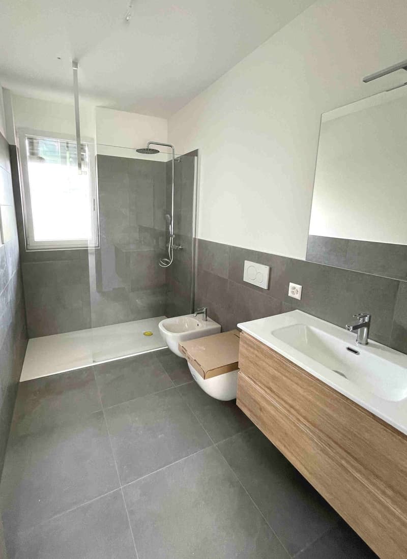 modern bathroom with gray tiles, glass shower enclosure, white sink, wooden cabinet, white toilet, bidet, power outlet