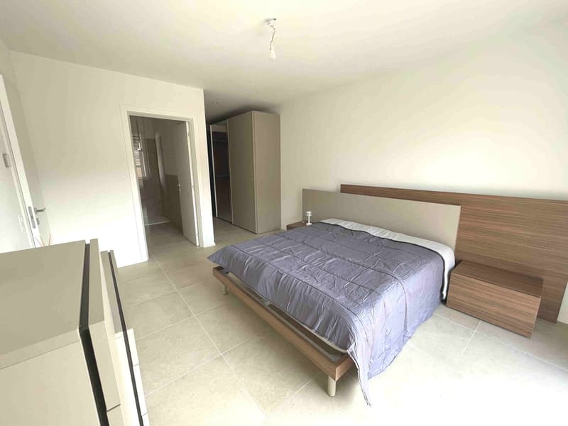 A bedroom with a bed, nightstand, lamp, and a wooden headboard. There is a wardrobe and an open door leading to another room. The room has white walls and tiled flooring.