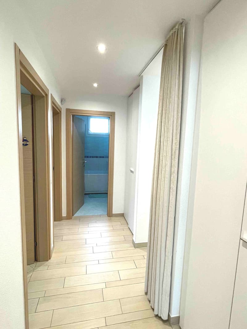 Empty hallway with wooden floors, white walls, and doors leading to different rooms, including a bathroom with a bathtub.