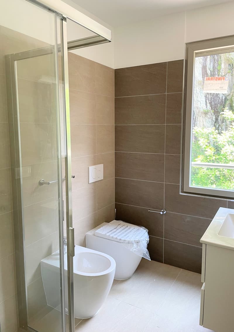 modern bathroom with white tiles, glass shower enclosure, white toilet, white sink, window with view