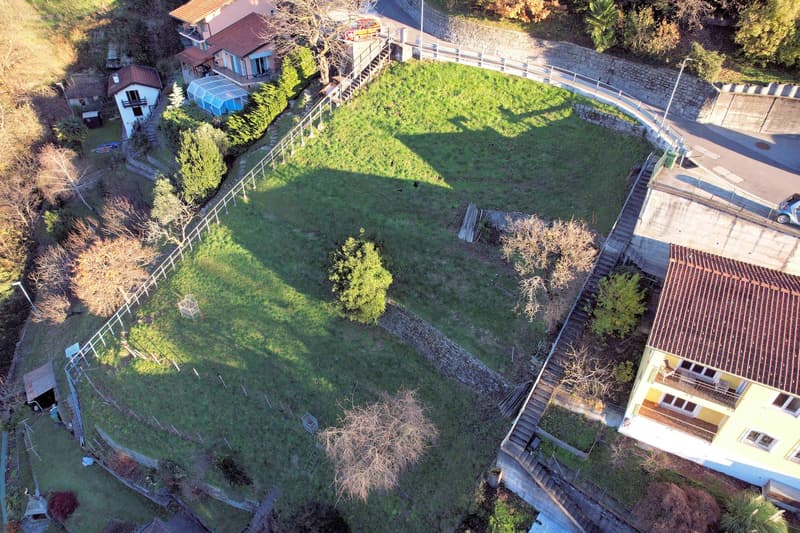Aerial view of a plot of land with a house and a building in the background, surrounded by trees and plants.