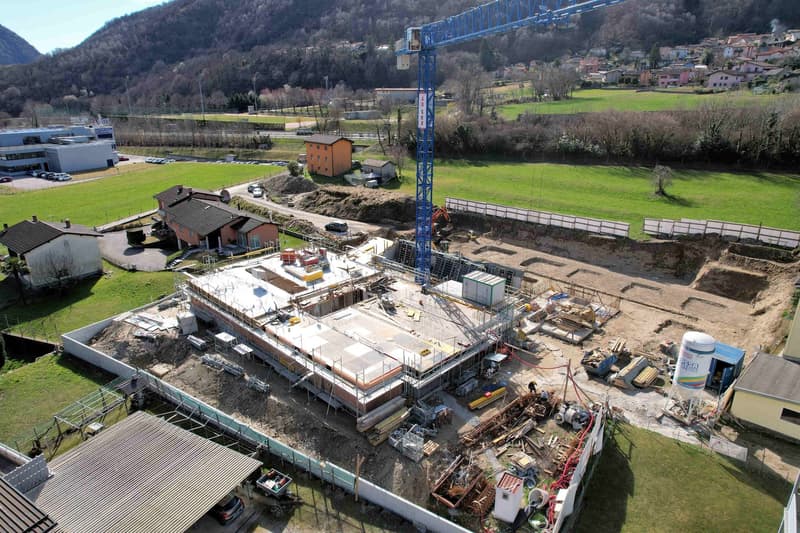 Under construction building, large crane, construction materials, surrounding houses, green fields, mountains in the background