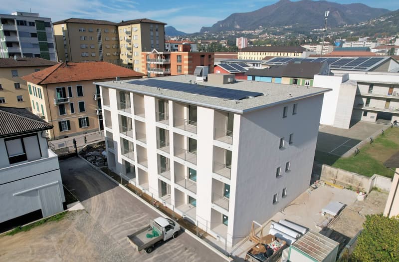 Aerial view of a modern apartment building with solar panels on the roof, balconies, and a parking area in front.