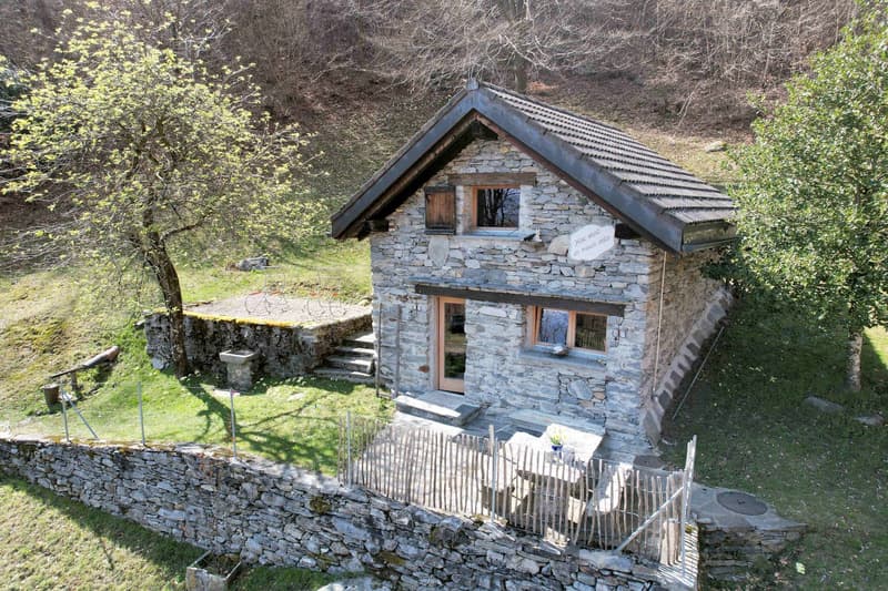 Stone house, wooden door, windows, surrounded by grass and trees, stone wall, wooden fence, table and chairs