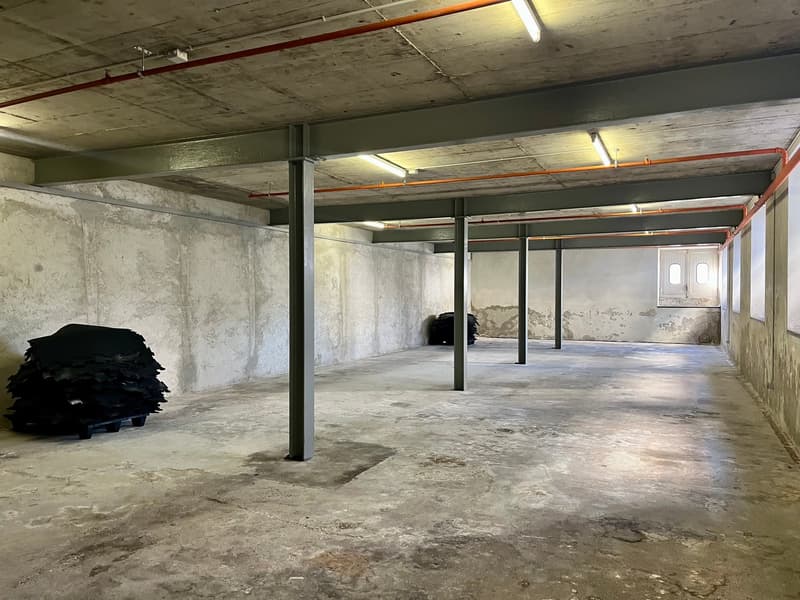 Open room, concrete floor, metal support beams, fluorescent lights on ceiling, doors, piles of materials