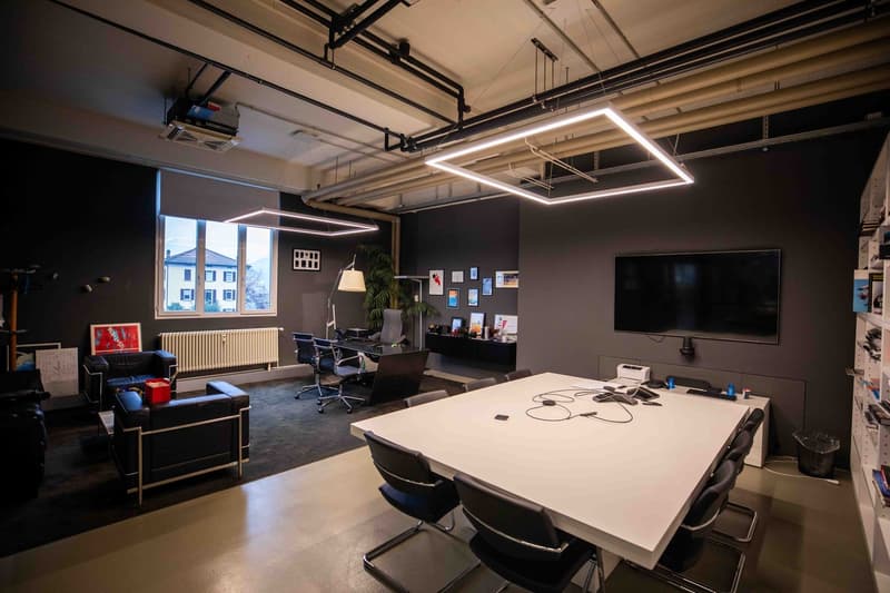 Modern furnished room with a large desk, modern furniture, TV mounted on the wall, windows, lamps, and a white shelf.