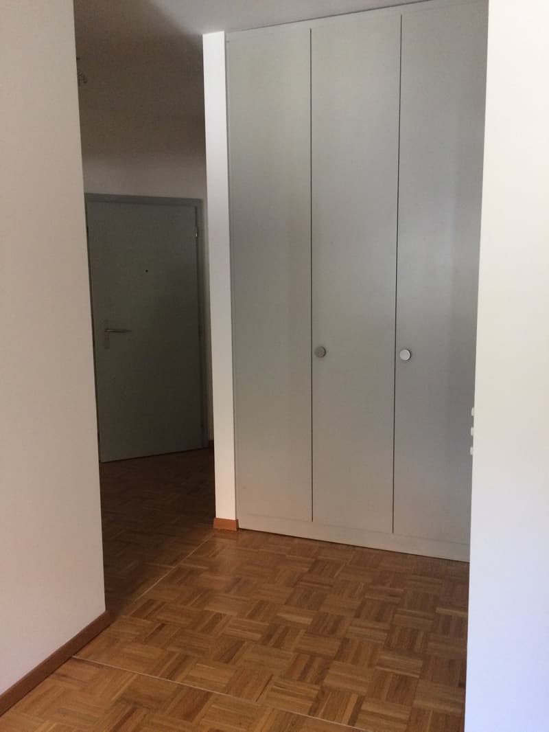 The image shows a bedroom with a large built-in wardrobe, a wooden parquet floor, and a door leading to another room.