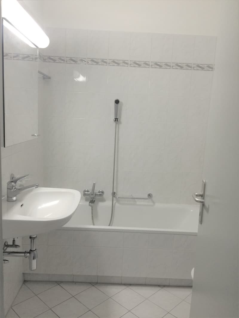The image shows a bathroom with white tiles on the walls and floor. There is a bathtub with a shower head, a sink, and some bathroom accessories such as towel racks.