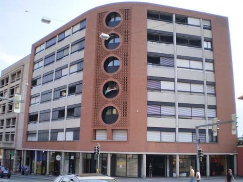A tall multistory building with a brick facade, large arched windows, and three circular patterns on the front, with people walking on the sidewalk in front