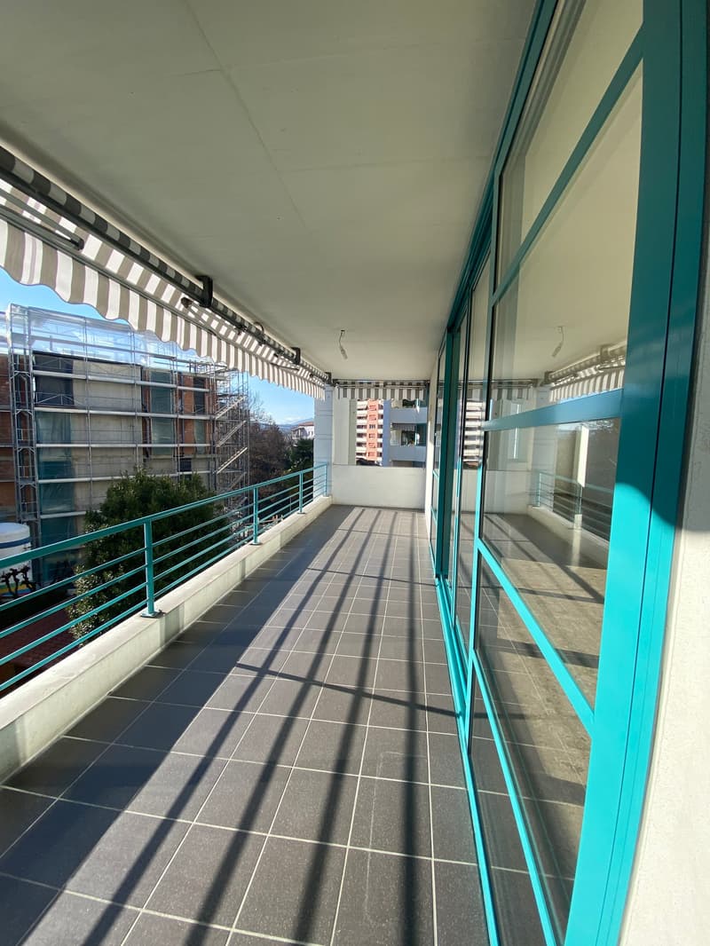 Modern balcony with teal railings, tiled floor, large windows, shaded area