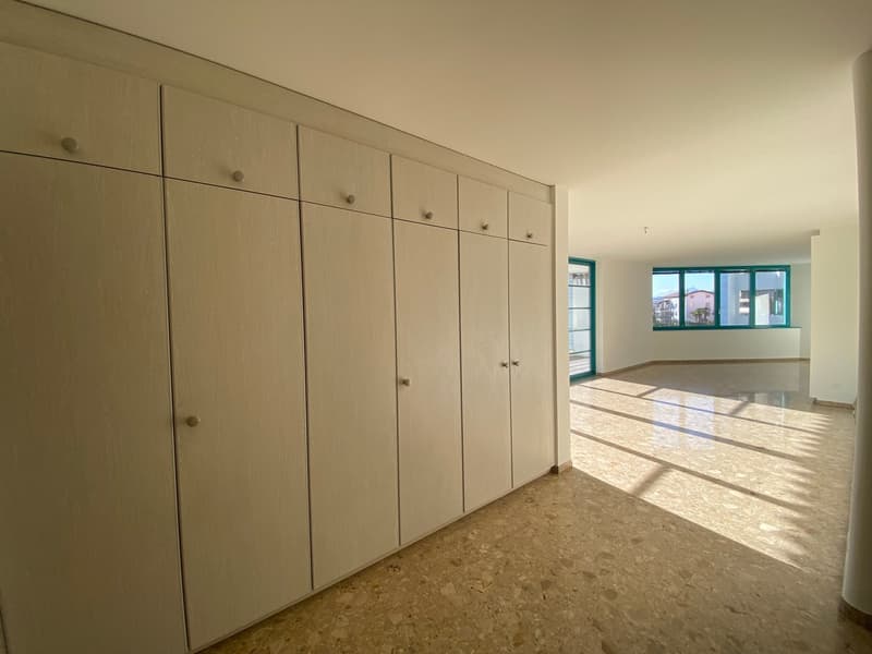 A hallway with white wooden built-in wardrobes and a large window with green frames leading to a balcony.