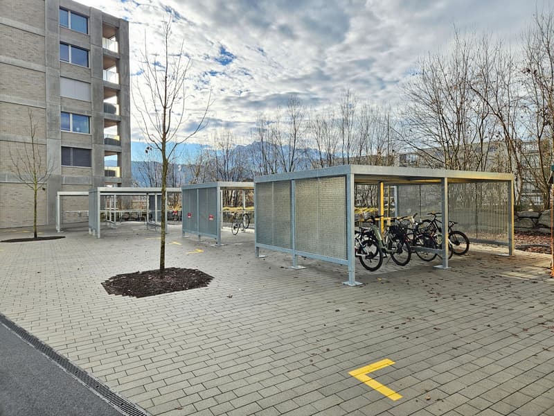 Bicycle parking shelter in urban setting, paved area, several bicycles parked, multi-story residential building in background, cloudy sky