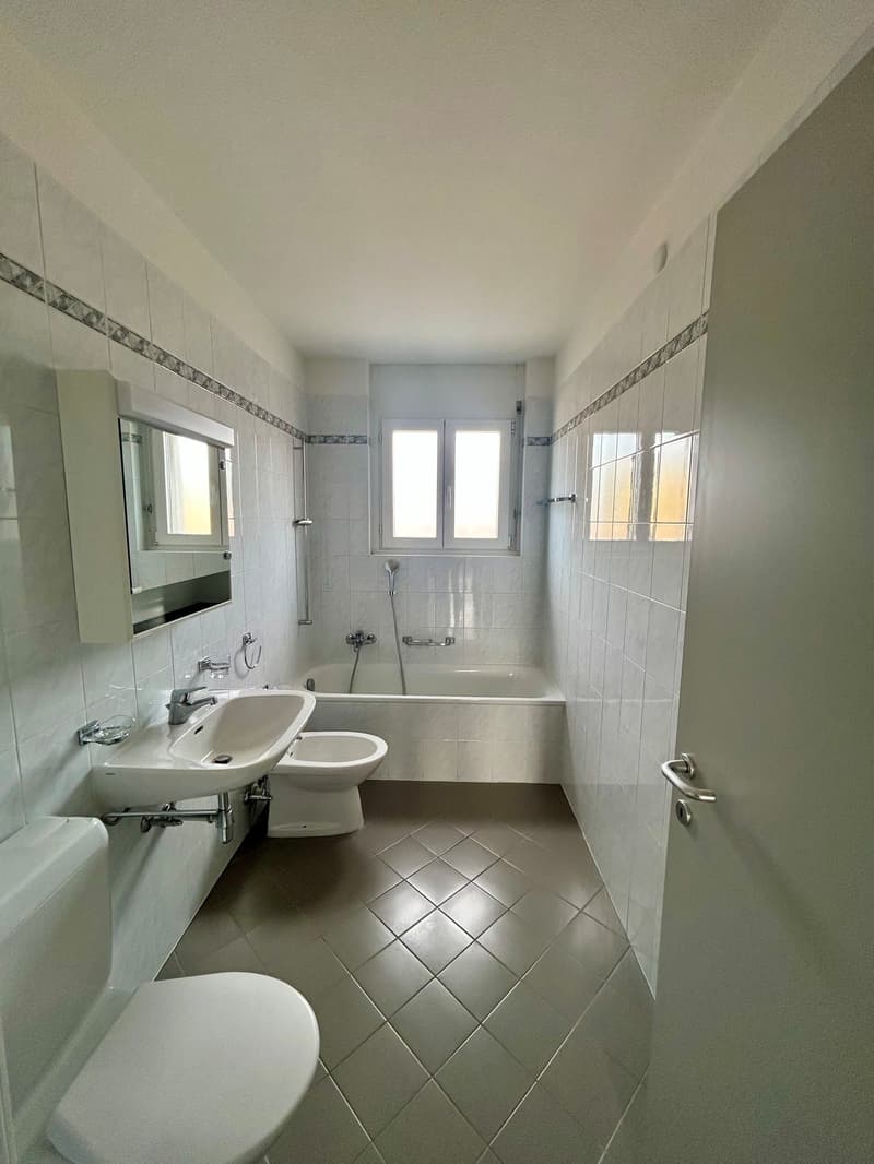 The image shows a spacious bathroom with white tiles on the walls and floor. It has a bathtub, a sink, and a toilet. The bathroom appears to be well-lit with a window providing natural light.