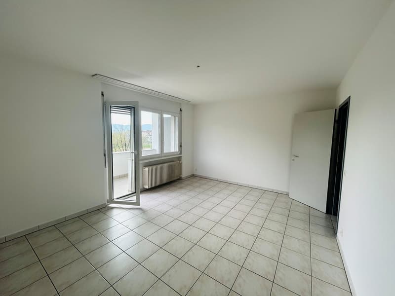 Spacious room with tile flooring, large windows, and a sliding glass door leading to a balcony or terrace. The room appears to be empty, with white walls and a radiator visible.